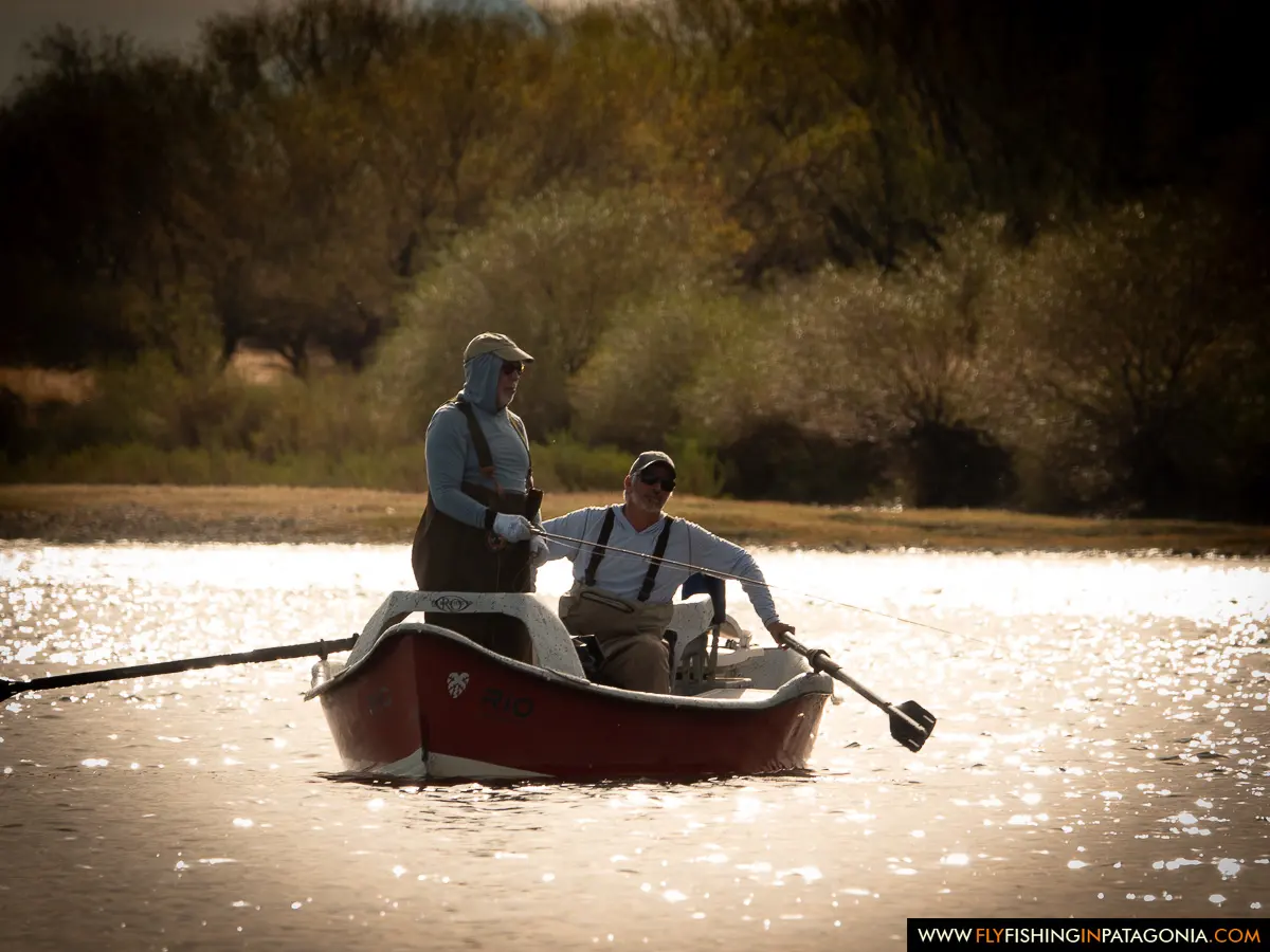 Anglers rowing a red drift boat through backlit water on the Collon Cura River